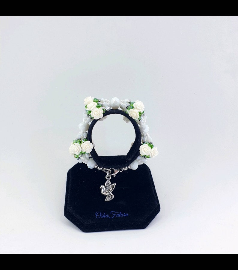 Puede incluir: Pulsera blanca de cuentas con flores blancas y hojas verdes. La pulsera tiene un dije de paloma plateada y el texto "Osha Futura" en un expositor de terciopelo negro.