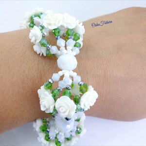 Puede incluir: Un brazalete blanco de cuentas con cuentas verdes y plateadas y detalles de flores blancas. El brazalete est&aacute; en la mu&ntilde;eca de una persona.