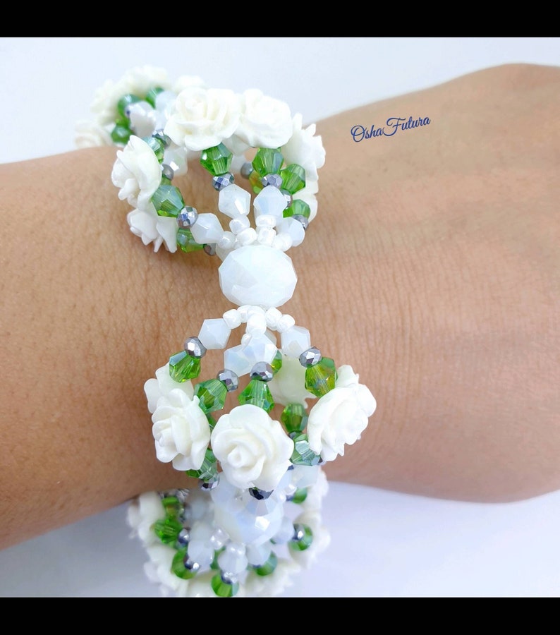 Puede incluir: Un brazalete blanco de cuentas con cuentas verdes y plateadas y detalles de flores blancas. El brazalete est&aacute; en la mu&ntilde;eca de una persona.