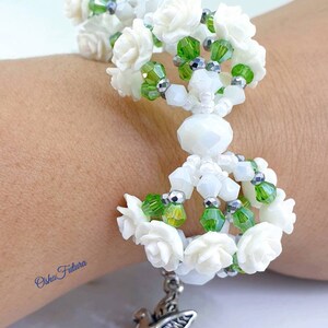 Puede incluir: Pulsera de cuentas blancas con cuentas verdes y plateadas, con dijes de flores blancas y un dije de paloma plateada. La pulsera est&aacute; en la mu&ntilde;eca de una persona.