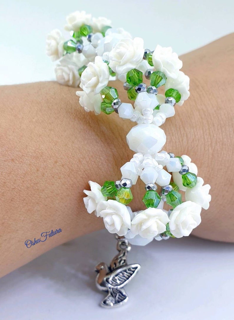 Puede incluir: Pulsera de cuentas blancas con cuentas verdes y plateadas, con dijes de flores blancas y un dije de paloma plateada. La pulsera est&aacute; en la mu&ntilde;eca de una persona.