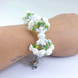 Puede incluir: Pulsera de cuentas blancas con detalles de flores blancas y cuentas verdes. La pulsera tiene un dije de paloma plateado.
