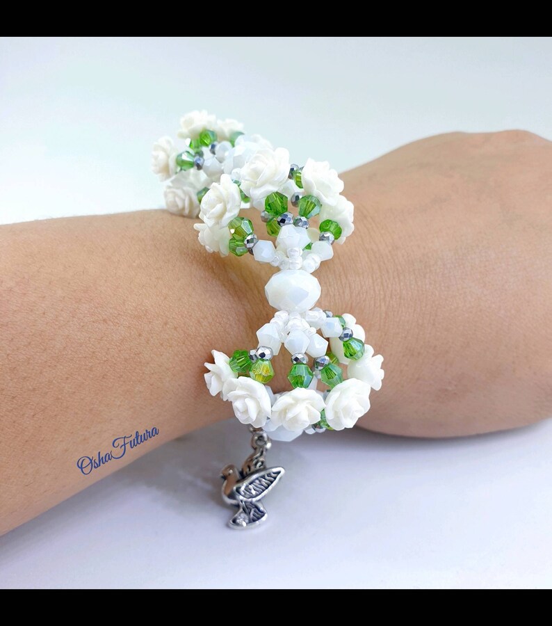 Puede incluir: Pulsera de cuentas blancas con detalles de flores blancas y cuentas verdes. La pulsera tiene un dije de paloma plateado.