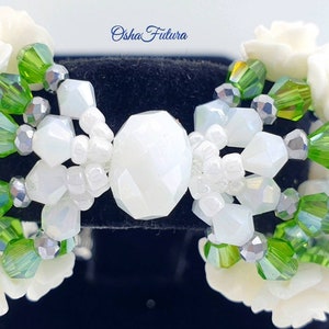 Puede incluir: Pulsera de cuentas blancas y verdes con detalles de flores blancas. La pulsera presenta una gran cuenta blanca facetada en el centro.