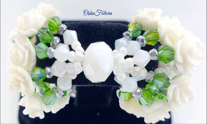 Puede incluir: Pulsera de cuentas blancas y verdes con detalles de flores blancas. La pulsera presenta una gran cuenta blanca facetada en el centro.