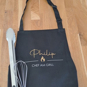 Könnte beinhalten: Schwarze Schürze mit dem Namen "Philip" in goldener Schrift, einer Flammen-Grafik und dem Text "CHEF AM GRILL" in Weiß. Eine Edelstahlzange und ein Schneebesen liegen neben der Schürze.