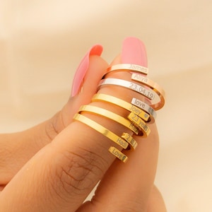 May include: A hand wearing a stack of gold and silver rings with engraved words, including "LOVE", "JAMES", "23.02.19", and "FOREVER".