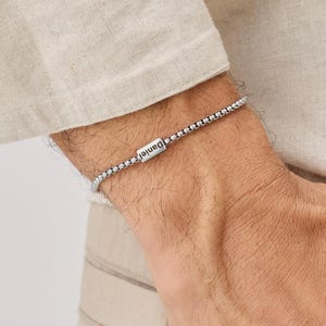 Könnte beinhalten: Ein silbernes Armband mit dem eingravierten Namen "Daniel" auf einem rechteckigen Balken. Das Armband besteht aus kleinen, runden Perlen und wird am Handgelenk getragen. Der Hintergrund ist eine Person, die ein helles Hemd trägt.