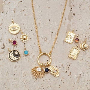 May include: Gold necklace with a variety of charms, including a hamsa hand, an evil eye, a crescent moon, a sun, and a star. The charms are attached to a gold chain with a circle pendant.