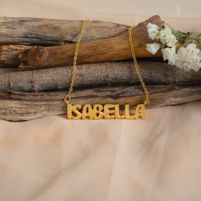 May include: A gold-colored necklace with the name "ISABELLA" in block letters. The necklace is displayed on a wooden surface with a beige background and white flowers.