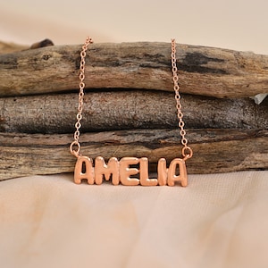 May include: A rose gold necklace with the name "AMELIA" in block letters. The pendant hangs from a delicate chain, displayed on a rustic wooden background. The necklace is a personalized jewelry item.