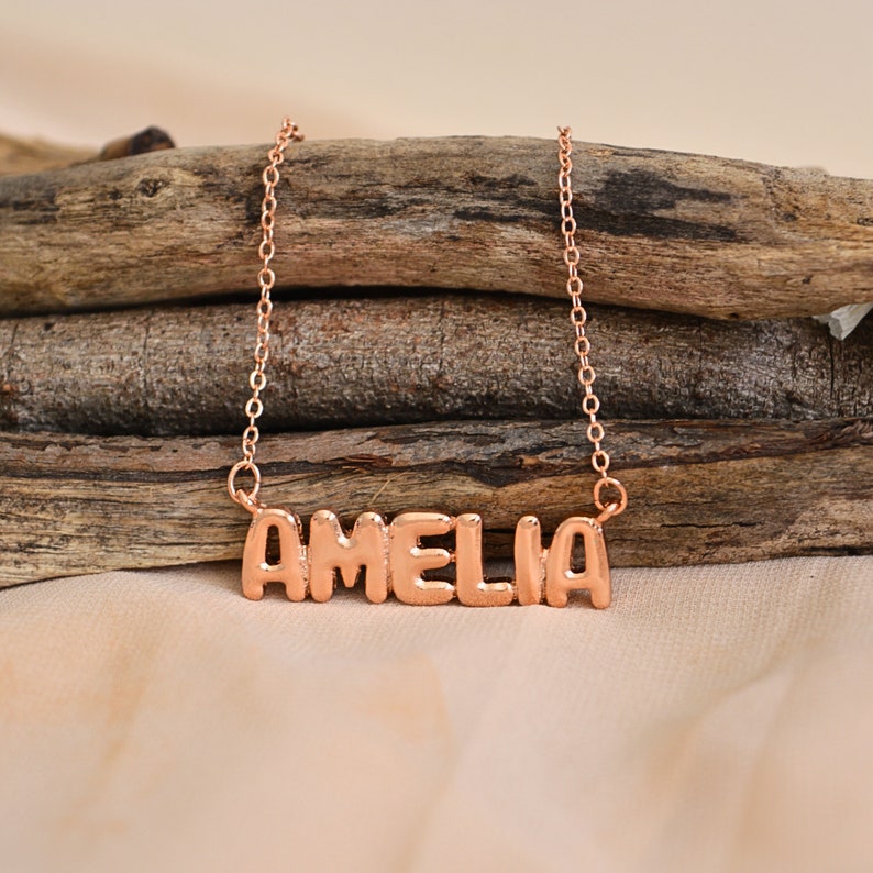 May include: A rose gold necklace with the name "AMELIA" in block letters. The pendant hangs from a delicate chain, displayed on a rustic wooden background. The necklace is a personalized jewelry item.