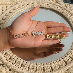 May include: Three name necklaces in rose gold, silver, and gold. The names "AMELIA", "EMMA", and "ISABELLA" are displayed in block letters. The necklaces are on delicate chains, presented on a hand against a neutral background.