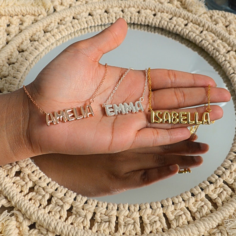 May include: Three name necklaces in rose gold, silver, and gold. The names "AMELIA", "EMMA", and "ISABELLA" are displayed in block letters. The necklaces are on delicate chains, presented on a hand against a neutral background.
