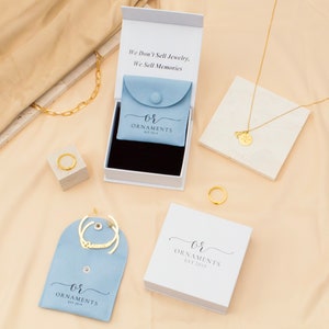 May include: An assortment of gold jewelry, including a chain necklace, ring, and pendant necklace, displayed with packaging. The packaging includes white boxes and light blue pouches, some with the text "We Don't Sell Jewelry, We Sell Memories" and "OR ORNAMENTS EST 2019".