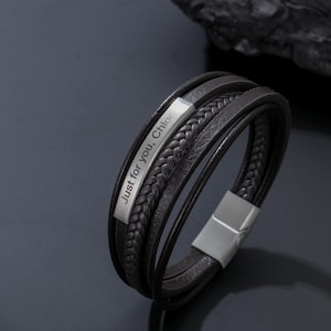 Könnte beinhalten: Schwarzes Lederarmband mit mehreren Strängen und einem silbernen Verschluss. Das Armband hat einen geflochtenen Lederstrang und einen silbernen Balken mit der Aufschrift "Just for you, Chloe".