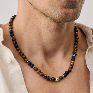 Puede incluir: Un collar de cuentas con cuentas de ojo de tigre y piedra de lava negra. El collar tiene una cuenta decorativa de color plateado en el centro. Las cuentas de ojo de tigre son marrones y doradas, y las cuentas de lava son negras.
