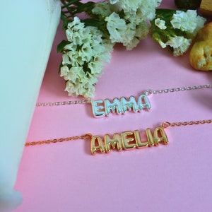 May include: Two name necklaces, one silver and one gold, are displayed on a pink surface. The silver necklace spells "EMMA", and the gold necklace spells "AMELIA". White flowers and a white object are in the background.