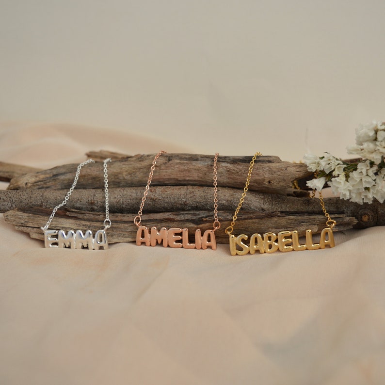 May include: Three name necklaces in silver, rose gold, and gold. The silver necklace spells "EMMA", the rose gold necklace spells "AMELIA", and the gold necklace spells "ISABELLA". Each necklace is on a delicate chain.
