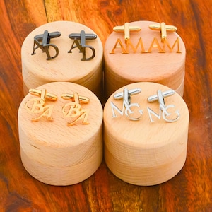 Op de afbeelding: Vier houten dozen met manchetknopen erin. De manchetknopen zijn goud, zilver en zwart. De dozen zijn gelabeld met de letters "AD", "AMAM", "BA" en "MG".