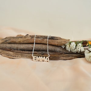 May include: A silver-colored necklace with the name "EMMA" spelled out in block letters. The necklace is displayed on a piece of driftwood, with white flowers and a beige background.