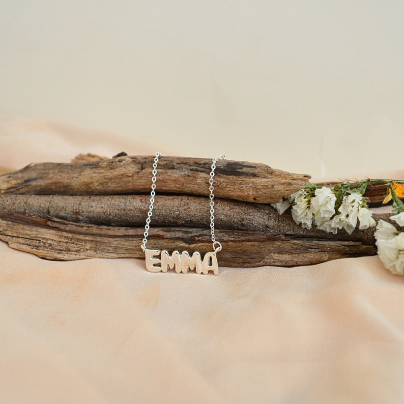 May include: A silver-colored necklace with the name "EMMA" spelled out in block letters. The necklace is displayed on a piece of driftwood, with white flowers and a beige background.