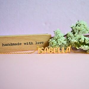 May include: A gold-colored necklace spelling "ISABELLA" in block letters. The necklace is on a delicate chain. A wooden block with the words "handmade with love" is in the background, along with white flowers, all set against a pink backdrop.