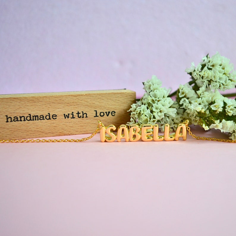May include: A gold-colored necklace spelling "ISABELLA" in block letters. The necklace is on a delicate chain. A wooden block with the words "handmade with love" is in the background, along with white flowers, all set against a pink backdrop.