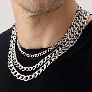 May include: A close-up of three silver chain necklaces of different thicknesses. The necklaces are layered, creating a bold look. The chains have a polished finish and are worn against a black top.