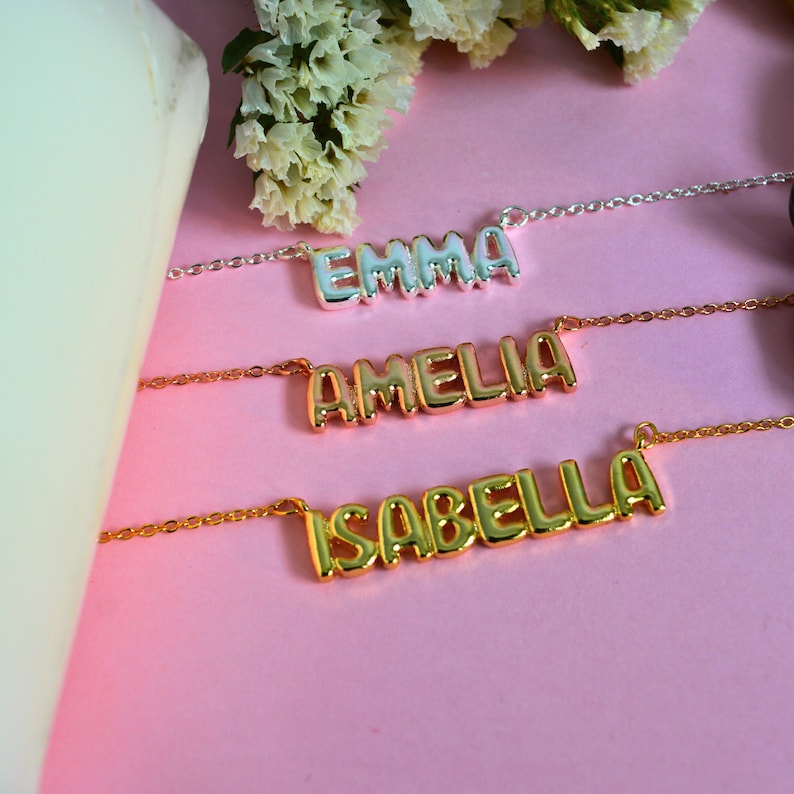 May include: Three personalized name necklaces on a pink background. The necklaces are gold and silver, with the names "EMMA", "AMELIA", and "ISABELLA" displayed in a bubble letter font. White flowers are in the background.