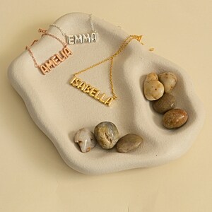 May include: Four name necklaces spelling "AMELIA", "EMMA", and "ISABELLA" in a block letter style. The necklaces are in rose gold, silver, and gold tones. The necklaces are displayed on a light gray, wavy-edged tray with several small, smooth stones.