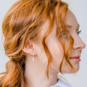 May include: A close-up of a gold hoop earring with a small, dangling letter "S" encrusted with small, clear crystals. The earring is worn on a person's ear, with the focus on the jewelry. The background is a soft, neutral color.
