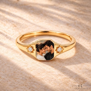 Op de afbeelding: Een goudkleurige zegelring met een rechthoekig middenpaneel waarop een foto van een omhelzend stel is afgedrukt. De ringband is aan beide zijden versierd met een kleine, stervormige zetting met daarin een enkele heldere, ronde steen.