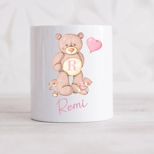May include: White ceramic mug featuring a brown teddy bear holding a pink heart balloon. Two smaller teddy bears are positioned on either side of the larger bear. The mug is inscribed with the name "Remi" in pink cursive writing.
