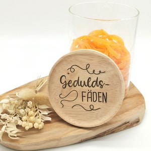 Könnte beinhalten: Ein Holzuntersetzer mit den Worten "Gedulds-Fäden" in dekorativer Schrift graviert. Der Untersetzer liegt auf einem Holzservierbrett mit Trockenblumen und einem Glasgefäß gefüllt mit orangefarbenen Süßigkeiten.