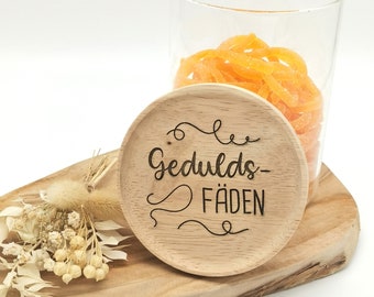 May include: A wooden coaster with the words "Gedulds-Fäden" engraved in a decorative font. The coaster sits on a wooden serving board with dried flowers and a glass jar filled with orange candy.