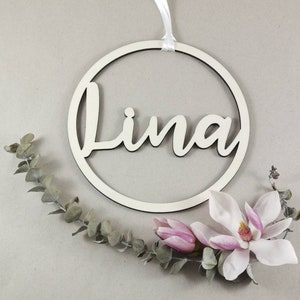 Könnte beinhalten: Ein weißer Holzkreis mit dem Namen "Lina" ausgeschnitten in einer kursivschrift. Der Kreis hängt an einem weißen Band und ist von einem Zweig Eukalyptus und rosa Magnolienblüten umgeben.