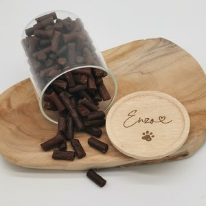 Könnte beinhalten: Ein Glasgefäß gefüllt mit braunen Hundefutter-Leckerlis kippt auf eine Holzoberfläche. Der Deckel des Gefäßes ist aus Holz und trägt den eingravierten Namen "Enzo" mit einem Herz und einem Pfotenabdruck.