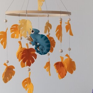 May include: A wooden baby mobile with a blue chameleon hanging from a branch. The mobile also features yellow and orange felt leaves.