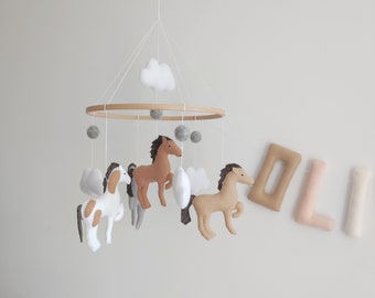 May include: A wooden mobile with felt horses and clouds hanging from a circular wooden frame. The mobile is suspended from the ceiling with a white string. The horses are brown, white, and gray. The clouds are white.