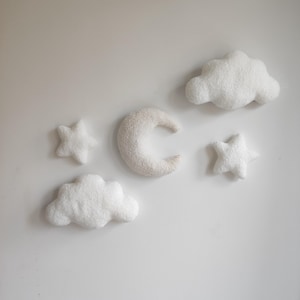 May include: A set of white felt wall decorations, including clouds, stars, and a crescent moon. The soft, plush shapes are arranged on a white background, creating a whimsical, celestial theme. The textured surfaces of the decorations add visual interest.