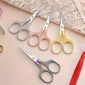 May include: Four pairs of small craft scissors with ornate floral engravings on the handles. The set includes one silver, one rose gold, one gold, and one iridescent multi-colored pair, each featuring sharp metal blades and a central pivot screw.