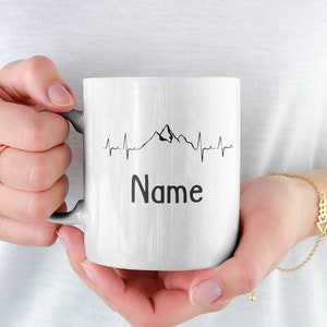 Könnte beinhalten: Weiße Keramiktasse mit einer schwarzen Bergkette und einer Herzschlaglinie. Der Text "Name" ist unter dem Motiv gedruckt.