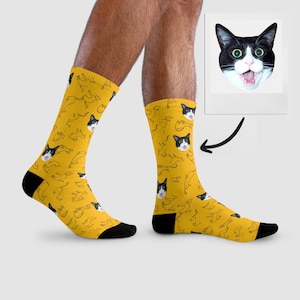 Könnte beinhalten: Ein Paar gelbe Socken mit einem sich wiederholenden Muster aus einem schwarzen und weißen Katzenkopf. Das Katzen Gesicht ist in jedem Muster gleich.
