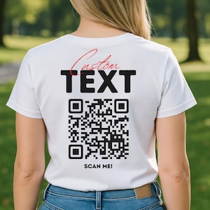 Puede incluir: Camiseta blanca con las palabras "Custom TEXT" en negro y rojo, y un código QR debajo. El texto "SCAN ME!" está impreso debajo del código QR. La persona lleva vaqueros azules.
