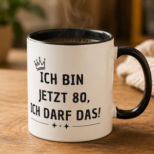Könnte beinhalten: Eine weiße Keramiktasse mit schwarzem Innenraum und Henkel, bedruckt mit einer schwarzen Krone und dem deutschen Text "ICH BIN JETZT 80, ICH DARF DAS!" auf der Vorderseite, ergänzt durch eine Zierlinie und Sternsymbole unter dem Text.