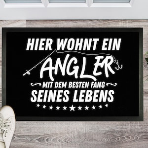 Könnte beinhalten: Schwarze Fußmatte mit weißem Text, der "Hier wohnt ein Angler mit dem besten Fang seines Lebens" (Hier wohnt ein Angler mit dem besten Fang seines Lebens) lautet. Der Text ist von Sternen umgeben.