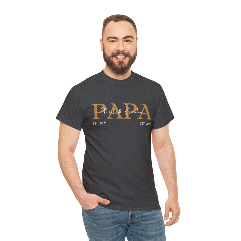 Puede incluir: Una camiseta gris oscuro jaspeada con la palabra "PAPA" impresa en dorado con el texto "Noah & Emilia" en escritura blanca debajo. Los textos "EST. 2021" y "EST. 2023" est&aacute;n impresos en blanco debajo de la palabra "PAPA".