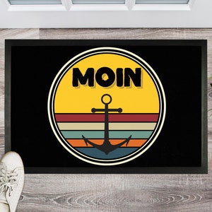 Può includere: Tappetino nero per porta con un cerchio giallo che contiene la parola "MOIN" in lettere nere e un'ancora nera su un tramonto in stile retrò.