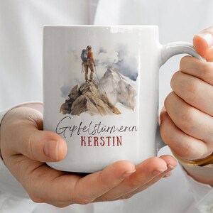 May include: White ceramic mug with a watercolor illustration of a hiker standing on a mountain peak. The text "Gipfelstürmerin KERSTIN" is printed on the mug.